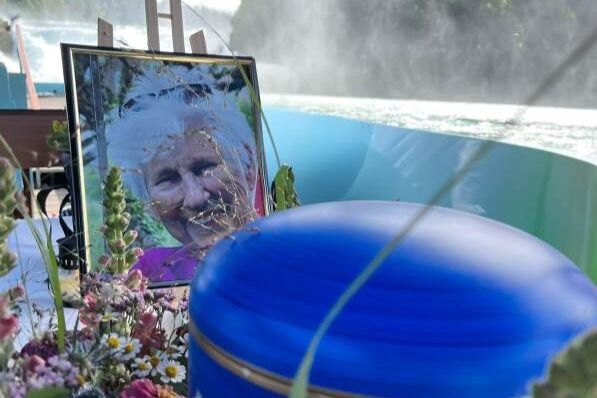 Eine blaue Urne im Vordergrund, links davon das Foto einer älteren verstorbenen Dame. Im Hintergrund erkennt man einen Fluss.