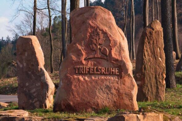 Drei nebeneinander stehende Steinfelsen (Hinkelsteine). Auf dem mittleren Stein ist das Wort "Trifelsruhe" eingemeißelt.
