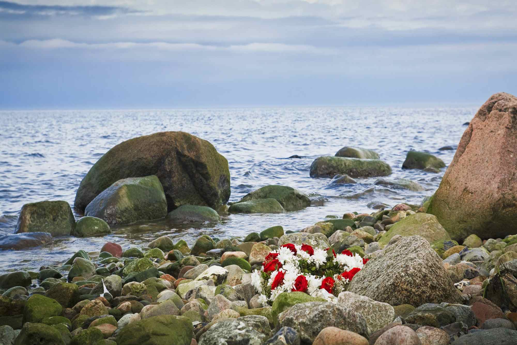 Im Vordergrund sind mehrere Steine und ein Blumenschmuck ist darauf gebettet. Im Hintergrund ist das Meer.