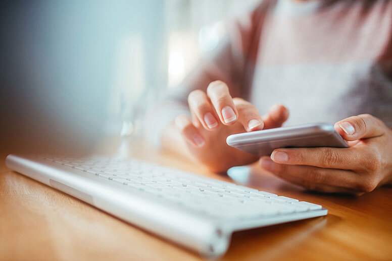 Auf einem Schreibtisch liegt eine Tastatur. Davor sitzt eine Person und hält ein Handy in der Hand.