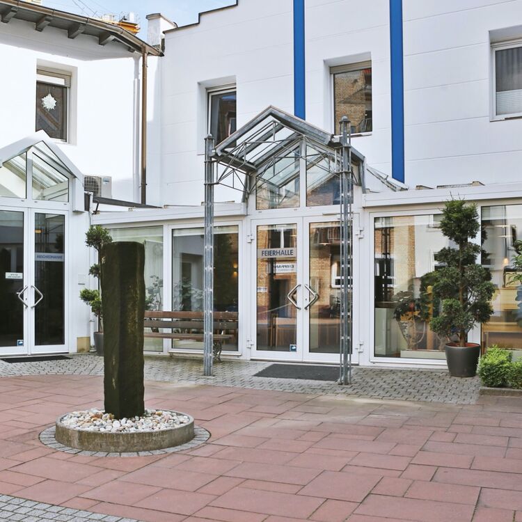 Eingang zur Feierhalle des Bestattungsinstituts mit Glasüberdachung und Steinbrunnen.